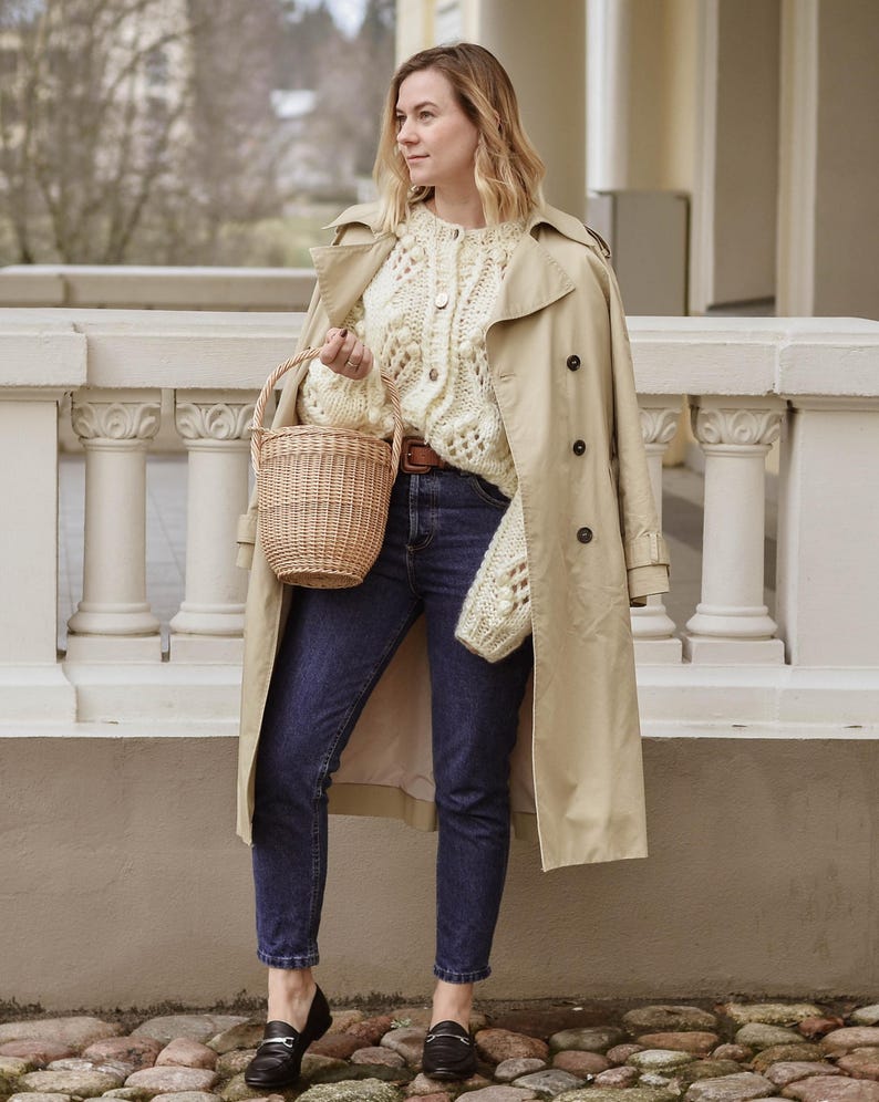 May include: A woman in a beige trench coat, cream-coloured knitted cardigan, and dark blue jeans, holding a woven basket. She wears black loafers. The background shows a white balustrade and a blurred outdoor scene.