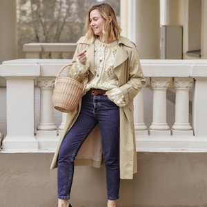 May include: A woman in a beige trench coat, cream knit cardigan, and dark blue jeans. She carries a wicker basket and wears black loafers. The outfit is a stylish combination for a casual, everyday look.