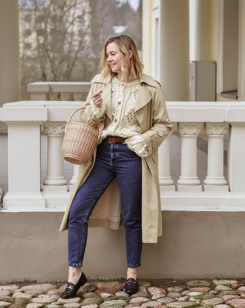 May include: A woman in a beige trench coat, cream knit cardigan, and dark blue jeans. She carries a wicker basket and wears black loafers. The outfit is a stylish combination for a casual, everyday look.