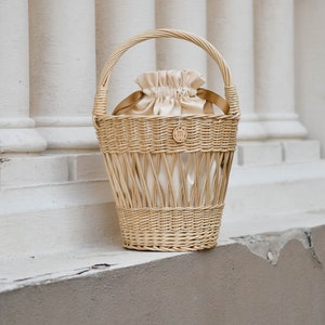 May include: A light brown wicker basket with a handle, holding a cream-colored drawstring bag. The basket has a woven design and a small wooden emblem. The background features white columns.
