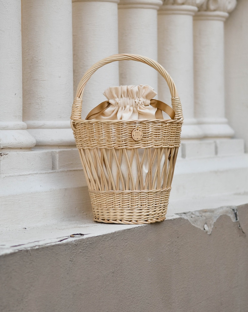 May include: A light brown wicker basket with a handle, holding a cream-colored drawstring bag. The basket has a woven design and a small wooden emblem. The background features white columns.