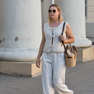 Pu&ograve; includere: Una donna indossa un gilet all'uncinetto color crema con chiusure a fiocco e pantaloni chiari. Porta una piccola borsa a cestino in vimini con cinturini in pelle. Indossa occhiali da sole e cammina su un marciapiede.