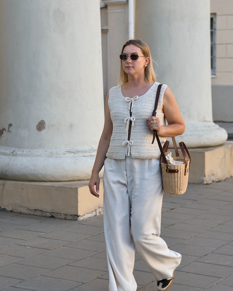 Pu&ograve; includere: Una donna indossa un gilet all'uncinetto color crema con chiusure a fiocco e pantaloni chiari. Porta una piccola borsa a cestino in vimini con cinturini in pelle. Indossa occhiali da sole e cammina su un marciapiede.