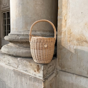 May include: A light brown woven wicker basket with a rounded body and a curved handle. The basket has a small, circular emblem on the front. The basket is sitting on a stone ledge next to a large column.