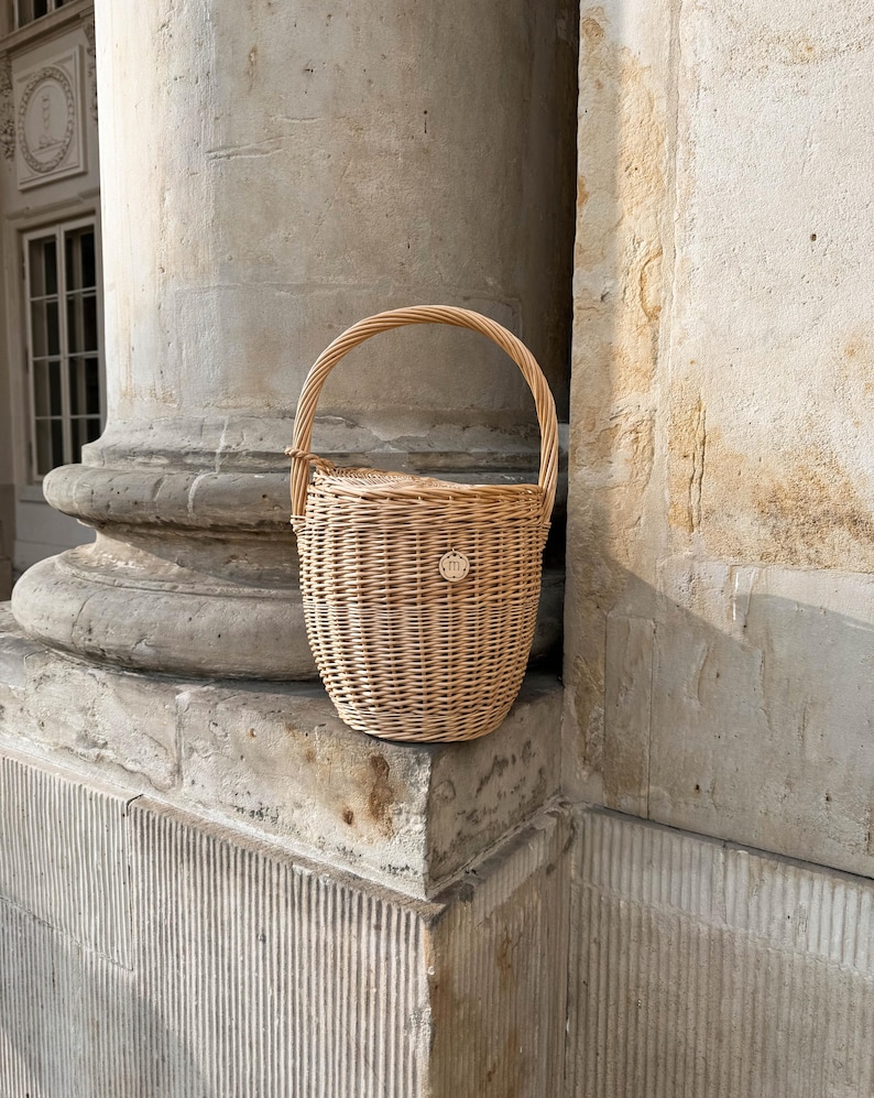 May include: A light brown woven wicker basket with a rounded body and a curved handle. The basket has a small, circular emblem on the front. The basket is sitting on a stone ledge next to a large column.