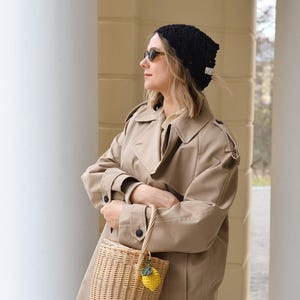 May include: A tan trench coat, black knit hat, and a woven basket bag with a yellow lemon charm. The woman is wearing sunglasses. The background features white columns and a light-colored wall.