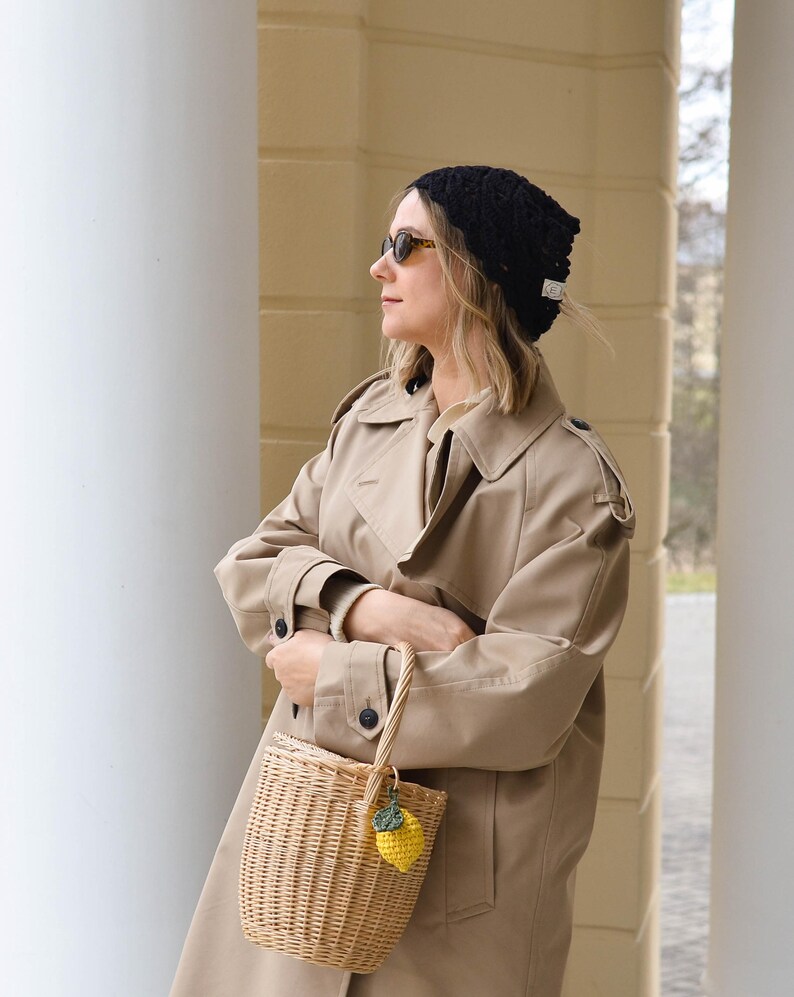 May include: A tan trench coat, black knit hat, and a woven basket bag with a yellow lemon charm. The woman is wearing sunglasses. The background features white columns and a light-colored wall.