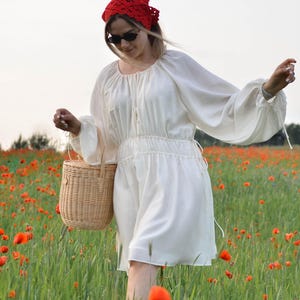 May include: A white dress with long sleeves and a gathered waist, paired with a red headscarf and sunglasses. A woven basket is carried in a field of green grass and red poppies. The dress has a round neckline and drawstring details.