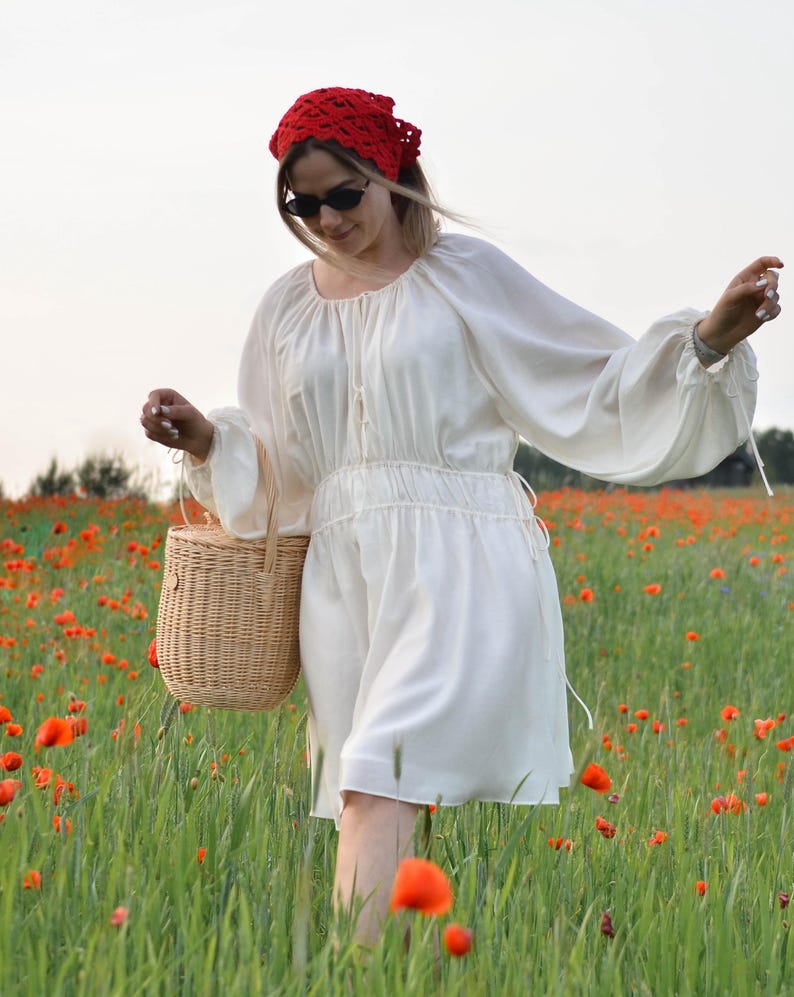 May include: A white dress with long sleeves and a gathered waist, paired with a red headscarf and sunglasses. A woven basket is carried in a field of green grass and red poppies. The dress has a round neckline and drawstring details.