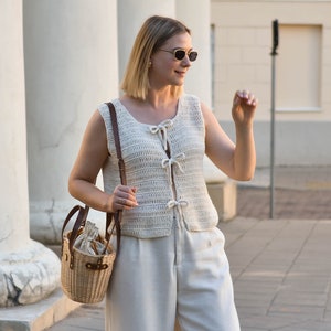 Pu&ograve; includere: Un gilet all'uncinetto color crema con chiusure a fiocco &egrave; abbinato a pantaloni coordinati. Una borsa a cestino intrecciata con un cinturino in pelle e una chiusura con coulisse &egrave; portata a tracolla. L'outfit &egrave; completato da occhiali da sole.