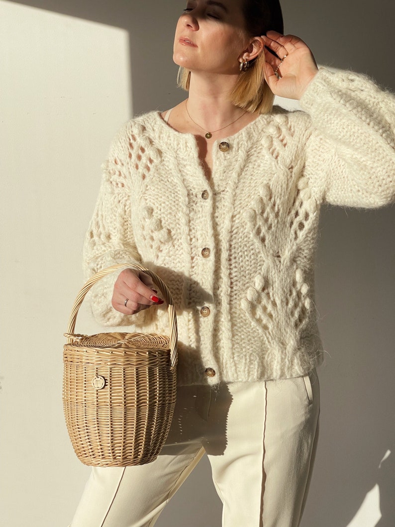 May include: Cream-coloured, open-knit cardigan with wooden buttons and a round neckline. The cardigan features textured bobbles and matching cream-coloured trousers. A light brown woven basket with a handle is held.