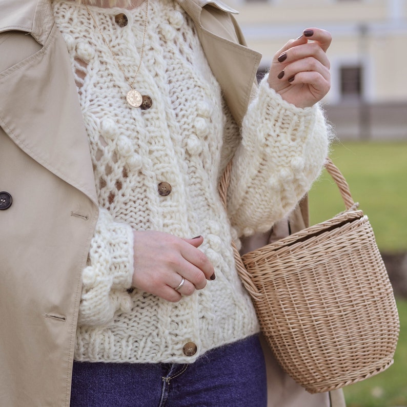 May include: Cream-colored, chunky knit cardigan with large wooden buttons. The cardigan features a textured pattern with bobbles and openwork details. A small, woven basket bag with a handle is also visible.