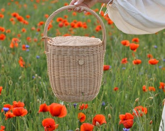 May include: A light brown wicker picnic basket with a rounded shape, a lid, and a handle. The basket is held in a field of red poppies and green grass.