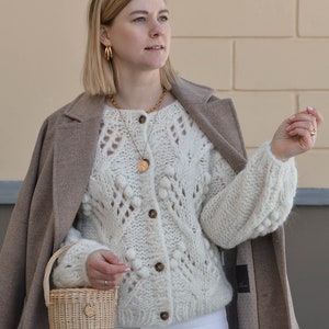 May include: A woman wearing a cream-coloured, chunky knit cardigan with a button-up front and a small woven basket bag. She is also wearing a gold necklace and earrings, a tan coat, and white trousers. The cardigan has a textured, open-knit design.