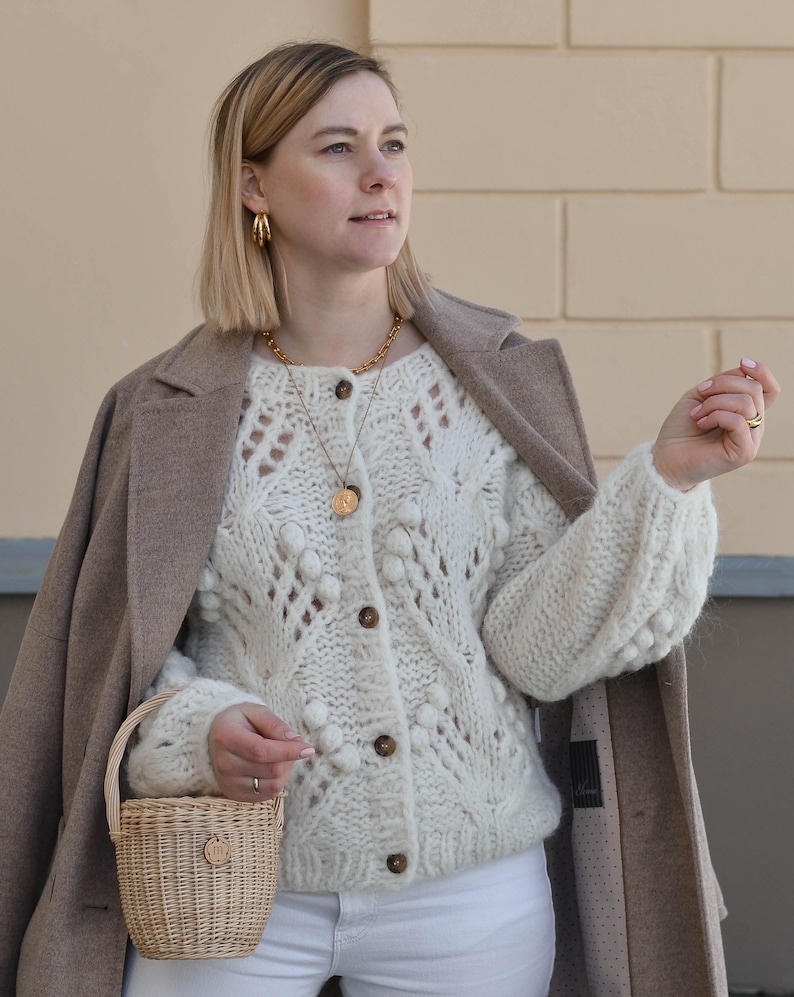 May include: A woman wearing a cream-coloured, chunky knit cardigan with a button-up front and a small woven basket bag. She is also wearing a gold necklace and earrings, a tan coat, and white trousers. The cardigan has a textured, open-knit design.