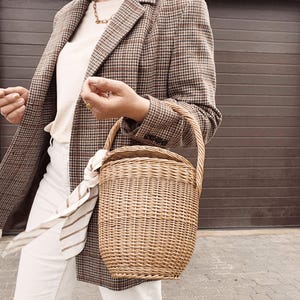 May include: A woven wicker basket with a curved handle is held in front of a person wearing a brown and black plaid blazer, a cream-colored shirt, and white pants. A striped scarf is tied to the basket handle.