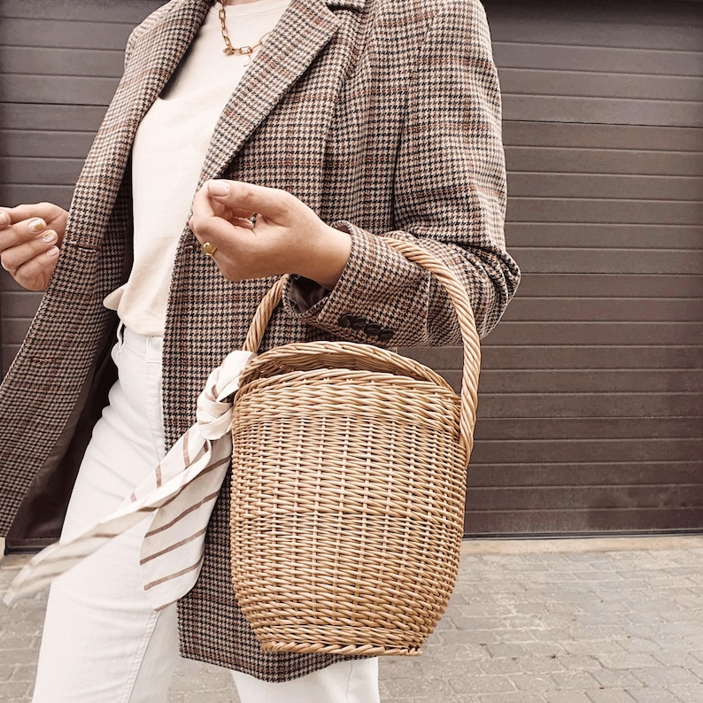 May include: A woven wicker basket with a curved handle is held in front of a person wearing a brown and black plaid blazer, a cream-colored shirt, and white pants. A striped scarf is tied to the basket handle.