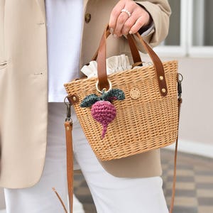 May include: Woven wicker handbag with brown leather handles and a detachable shoulder strap. The bag features a decorative crocheted beet charm in shades of purple and green. A small gold-colored charm with the letter "m" is attached to the bag.