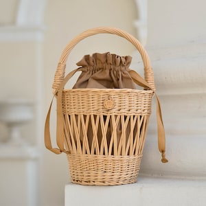 May include: Woven wicker basket with a tan drawstring interior and a matching handle. The basket has a round base and a woven design with tan leather straps. A small, round tag with an "m" is attached to the front.