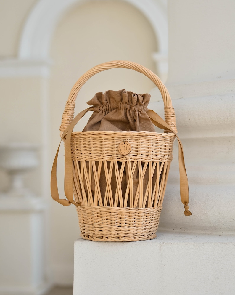 May include: Woven wicker basket with a tan drawstring interior and a matching handle. The basket has a round base and a woven design with tan leather straps. A small, round tag with an "m" is attached to the front.