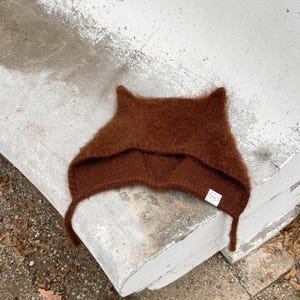 May include: A brown fuzzy knit hat with cat ears and a small white tag that says "COO" on it.