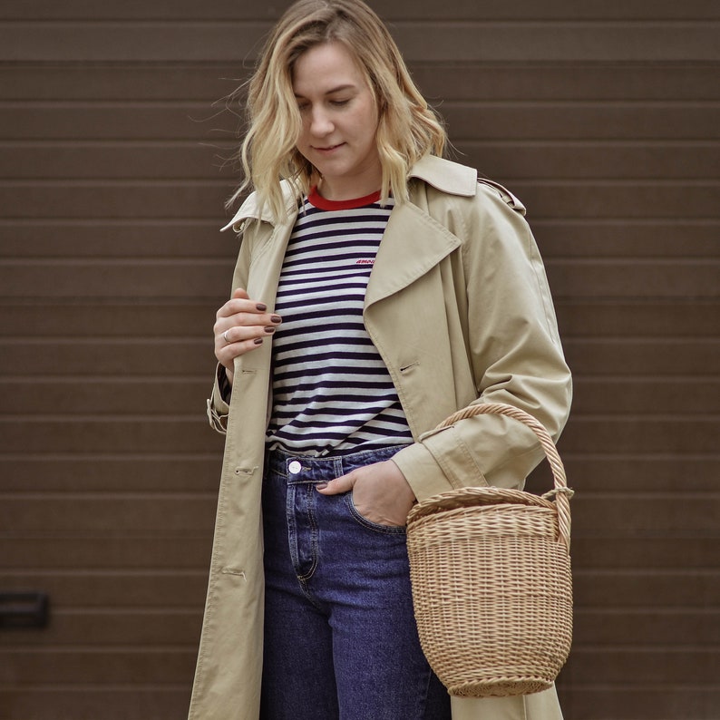 May include: A beige trench coat, striped top, and blue jeans. A woven basket bag with a handle is held. The top has a red collar and small text. The background is a brown garage door. The bag is made of woven wicker.