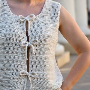 May include: A crocheted sleeveless top with a scoop neck and a tie detail at the front. The top is made in a light beige color.