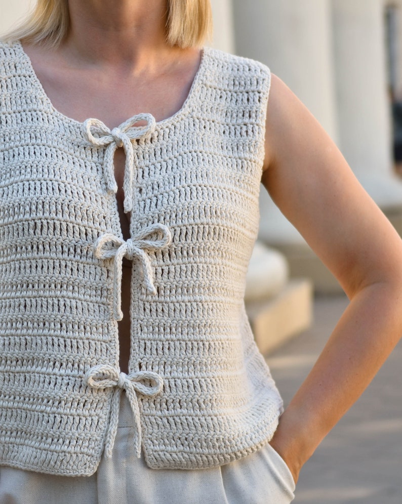 May include: A crocheted sleeveless top with a scoop neck and a tie detail at the front. The top is made in a light beige color.