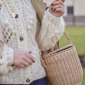 May include: Cream-coloured, open-weave cardigan with wooden buttons and a round, woven basket bag. The cardigan has a textured, bobble-stitch design. A gold-coloured pendant necklace is visible. The bag has a woven handle and a natural, light brown colour.