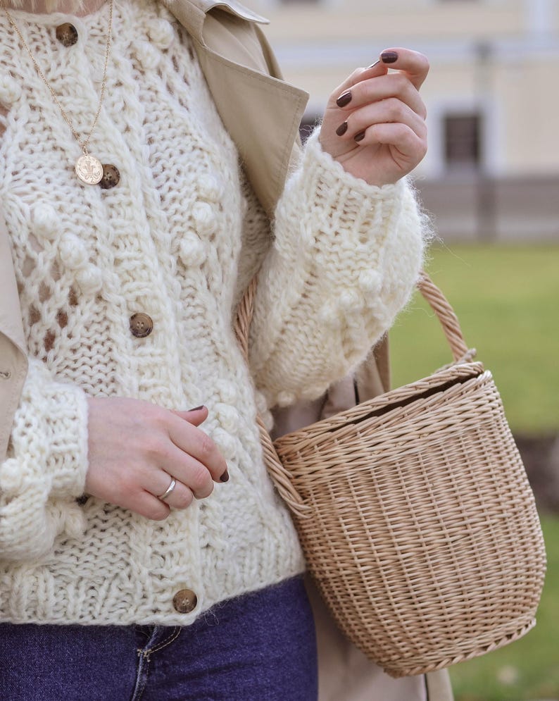 May include: Cream-coloured, open-weave cardigan with wooden buttons and a round, woven basket bag. The cardigan has a textured, bobble-stitch design. A gold-coloured pendant necklace is visible. The bag has a woven handle and a natural, light brown colour.