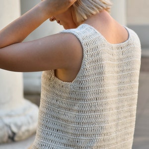 May include: A close-up of a crocheted sleeveless top in a cream color. The top has a loose, open weave and a scoop neck.