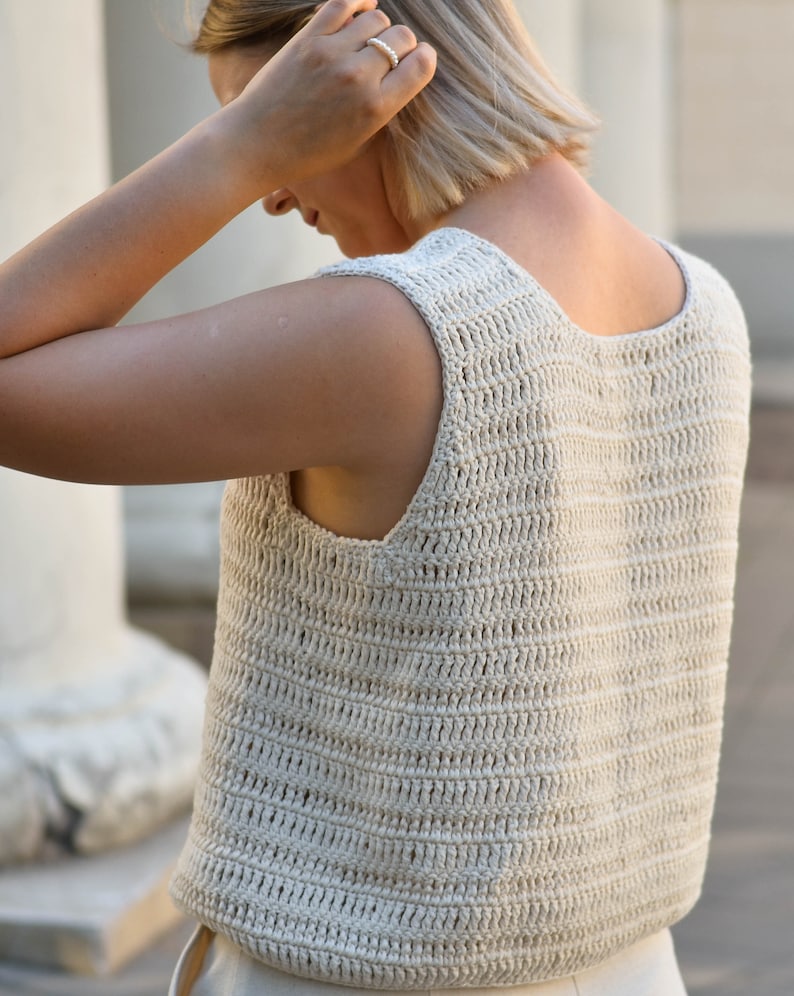 May include: A close-up of a crocheted sleeveless top in a cream color. The top has a loose, open weave and a scoop neck.