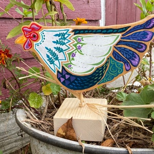 May include: A wooden chicken figurine painted in bright colors, including white, blue, green, and purple. The chicken is standing on a wooden base with straw around it.