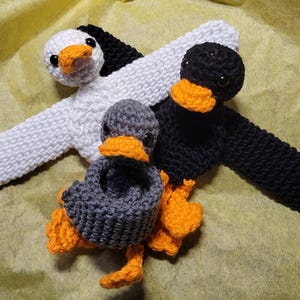 May include: Three crocheted duck plushies with orange beaks and feet. One duck is white, one is black, and one is gray.