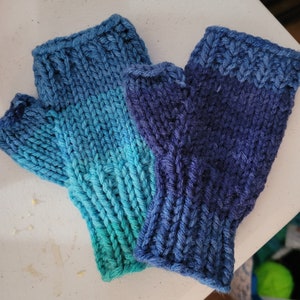 May include: A pair of knitted fingerless gloves in blue and teal ombre colors. The gloves are made with a simple knit stitch and have a textured look.