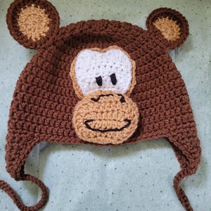 May include: Brown crocheted monkey hat with brown ears and a white face. The hat has two long ties.