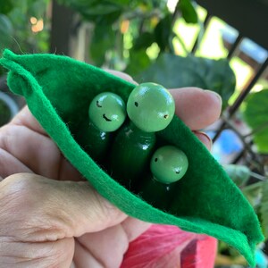 May include: A whimsical felt pea pod with three smiling green peas. The peas have black painted eyes and mouths. The pea pod is a vibrant green color, and the peas are a lighter shade of green.