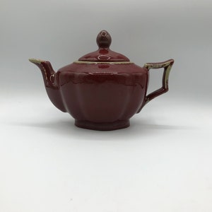Peut inclure: Une petite théière rouge foncé avec un couvercle et une anse. La théière a un corps arrondi et un bec légèrement évasé.