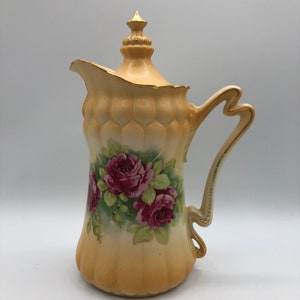 May include: A yellow ceramic jug with a lid and a floral design. The jug has a gold trim and is decorated with pink roses and green leaves.