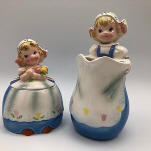 May include: A vintage ceramic sugar and creamer set featuring two Dutch girl figurines. The creamer is a jug with a blue and white floral design. The sugar bowl has a lid with a blue and white floral design. Both figurines are wearing blue and white dresses with pink and yellow flowers.