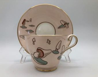 May include: A pink teacup and saucer set with a mushroom design. The cup and saucer are decorated with brown, green, and gold accents.