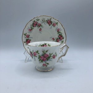 May include: A white teacup and saucer set with a pink rose floral pattern. The cup has a gold rim and two handles. The saucer has a gold rim and a scalloped edge.