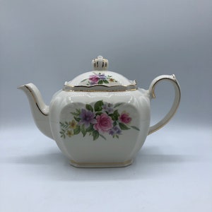May include: A white teapot with a floral design. The teapot has a square body with a gold trim and a lid with a knob. The floral design features pink, purple, and yellow flowers.