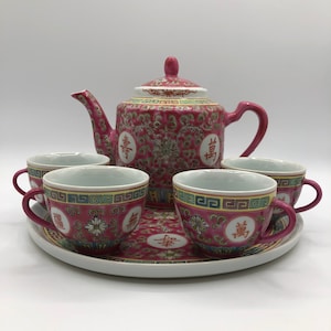 Peut inclure: Un service à thé chinois rose et blanc avec une théière et quatre tasses. L'ensemble présente un motif floral avec des caractères chinois et une bordure dorée. Les tasses et la théière ont un design assorti et sont sur un plateau assorti.