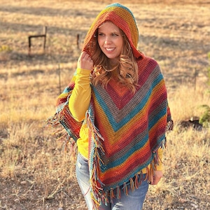May include: A woman wearing a colourful crocheted poncho with a hood and fringe. The poncho is made in shades of orange, red, brown, teal, and yellow. The woman is wearing blue jeans and a yellow shirt.