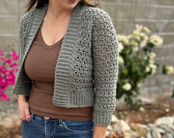 May include: A gray crochet cardigan with a cropped design. The open-front sweater has three-quarter sleeves and a textured stitch pattern. The model is wearing a brown V-neck top and blue jeans. The website address "hookedonhomemademahappiness.com" is visible.