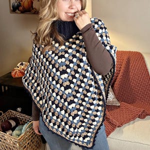 Puede incluir: Un poncho de ganchillo hecho a mano con un patrón de granny square. El poncho presenta una combinación de colores azul marino, blanco, beige y azul claro. El texto "hookedonhomemadehappiness.com" es visible en la esquina superior derecha.