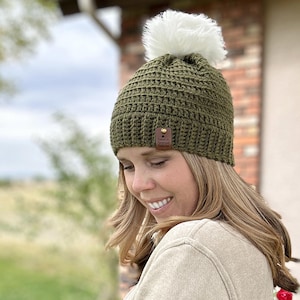 May include: Olive green crochet beanie with a fluffy white pom-pom on top. The hat has a small leather tag. The text "hooked on homemade happiness" is at the bottom of the image. A woman is wearing the hat.