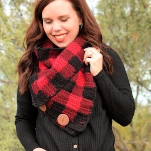 May include: A red and black plaid crochet infinity scarf with wooden buttons. The scarf is worn by a person with long brown hair and is styled with a black shirt and blue jeans.