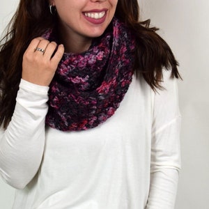 May include: A close-up of a woman wearing a white long-sleeved shirt and blue jeans. She is wearing a crocheted infinity scarf in shades of pink, purple, and gray.